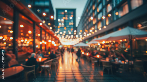 Blurred image shows bokeh of lights of restaurants on both sides of a street. A promenade in a city. Unknown people sit at tables by candlelight. In the background is a skyscraper.