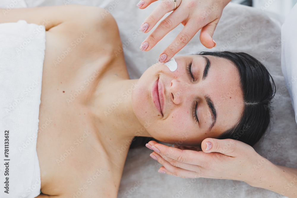The face of a young woman to whom a beautician applies cream to her skin. Facial. Facial skin care procedure. Moisturizing the face The face of a young woman to whom a beautician applies cream to her skin. Facial. Facial skin care procedure. Moisturizing the face