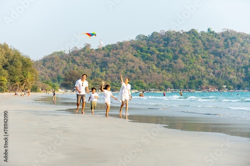 Happy Asian family travel nature ocean on summer beach holiday vacation. Parents and little child kid enjoy and fun outdoor activity lifestyle playing kite together on tropical island beach at sunset.