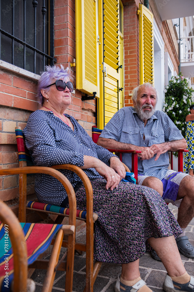 An elderly couple sits on a colorful chair near a brick wall of a house with yellow shutters