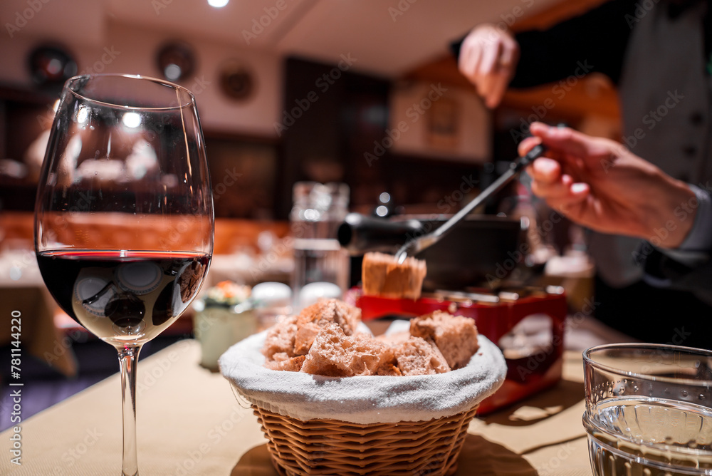 Naklejka premium Intimate dining scene with red wine, bread basket, fondue pot, person dipping food. Cozy ambiance, warm lighting. Possible location Europe or North America.