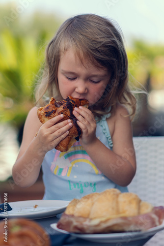 A beautiful little girl eats a croissant with chocolate with appetite