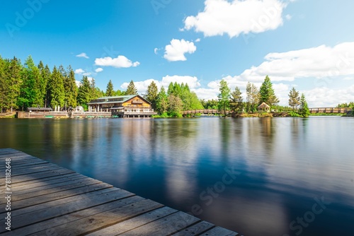 Smooth surface of the lake with green trees and a wooden building on the shore.