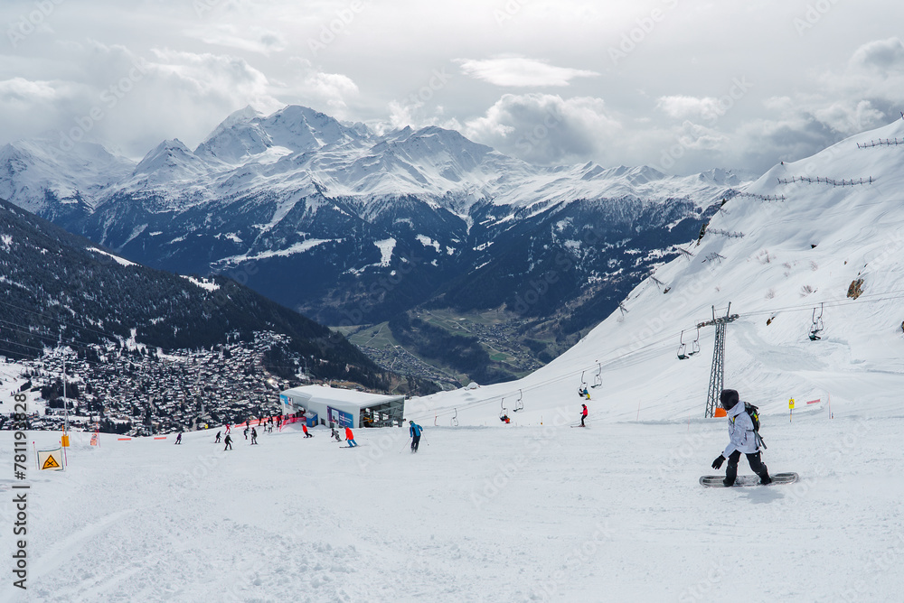 Snowy mountain landscape with winter sports activity. Snowboarders, skiers, chalets in alpine village. Majestic peaks, ski lift system, recreational scene. In a popular ski resort destination.