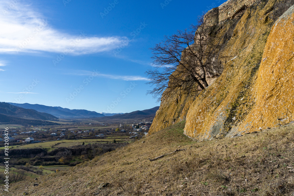 View of the village and mountains. The rock is covered with yellow lichen. Tree without leaves.  Bright blue sky with clouds.  Kveshi fortress. Georgia