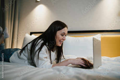Joyful woman lying on in bed and typing on her laptop, engaging in social media or remote work. 
