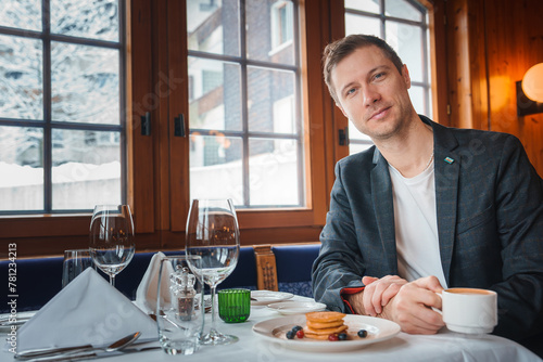 Elegant man dining in cozy restaurant with snowy view in possible ski resort, Zermatt. Smart attire, pancakes, coffee, smartphone, luxurious ambiance. photo