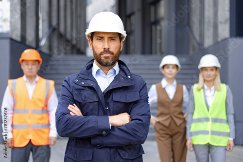 Portrait of serious young male engineer, owner in uniform and hard hat standing in front of camera with arms crossed on chest, behind is an interracial group of colleagues and contractors. © Liubomir