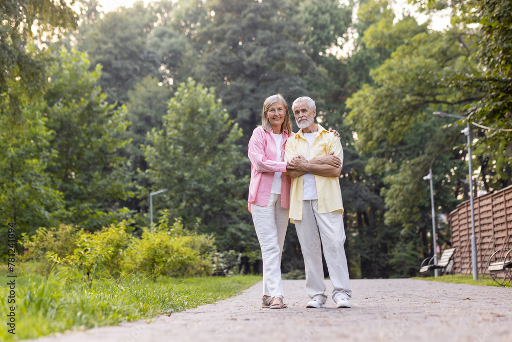 Portrait of senior gray-haired couple, man and woman standing in casual clothes in park, hugging each other and looking at camera. Portrait of senior gray-haired couple, man and woman standing in casual clothes in park, hugging each other and looking at camera.