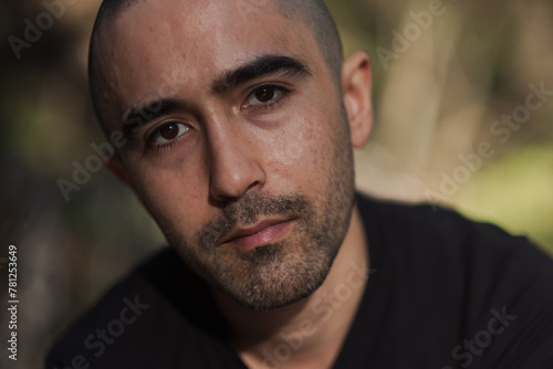 A man looks tilts his head to look at the camera for a direct portrait. Natural lighting streams in from the side, and the background is defocused. He has dense stubble and thick, dark eyebrows. photo