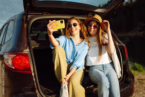Selfie while traveling. Happy women sits in the trunk of a car and uses a smartphone. The concept of traveling by car, active lifestyle, blogging.