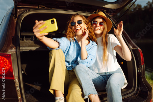 Selfie while traveling. Happy women sits in the trunk of a car and uses a smartphone. The concept of traveling by car, active lifestyle, blogging. © maxbelchenko