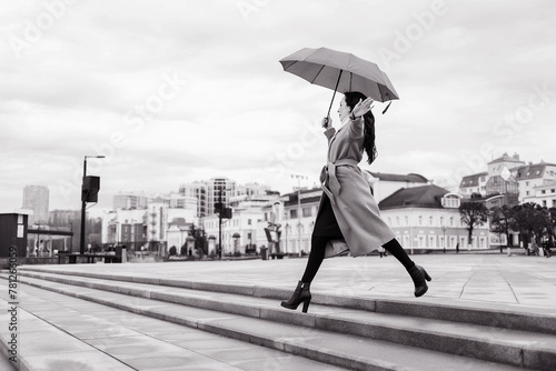 A girl in a light brown coat and a light scarf under an umbrella in a jump shot on a cloudy April evening against the background of Cheboksary photo