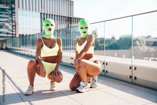 Two beautiful sexy women in green underwear. Models wearing bandit balaclava mask. Hot seductive female in nice lingerie posing in the street at sunny day, blue sky. Crime and violence. Perfect body