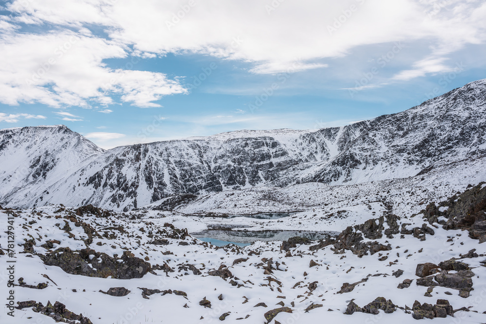 Cold snow-white landscape of snowy valley with alpine lake against snow-covered rocky mountain range under clouds in blue sky. Glacial lake, pure white snow and stones in high mountains in sunny day.