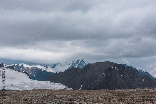 Dramatic misty top view from stone pass to big glacier tongue among sharp rocks and large snow-capped pinnacle in rainy low clouds. Dark atmospheric mountain silhouettes in rain in gray cloudy sky.