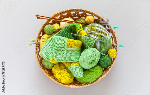 Top view of wicker basket with needlework: balls and skeins of green and yellow yarn, knitting needles and striped handmade knitted fabric. DIY concept. Flat lay, copy space, top view, closeup, mockup