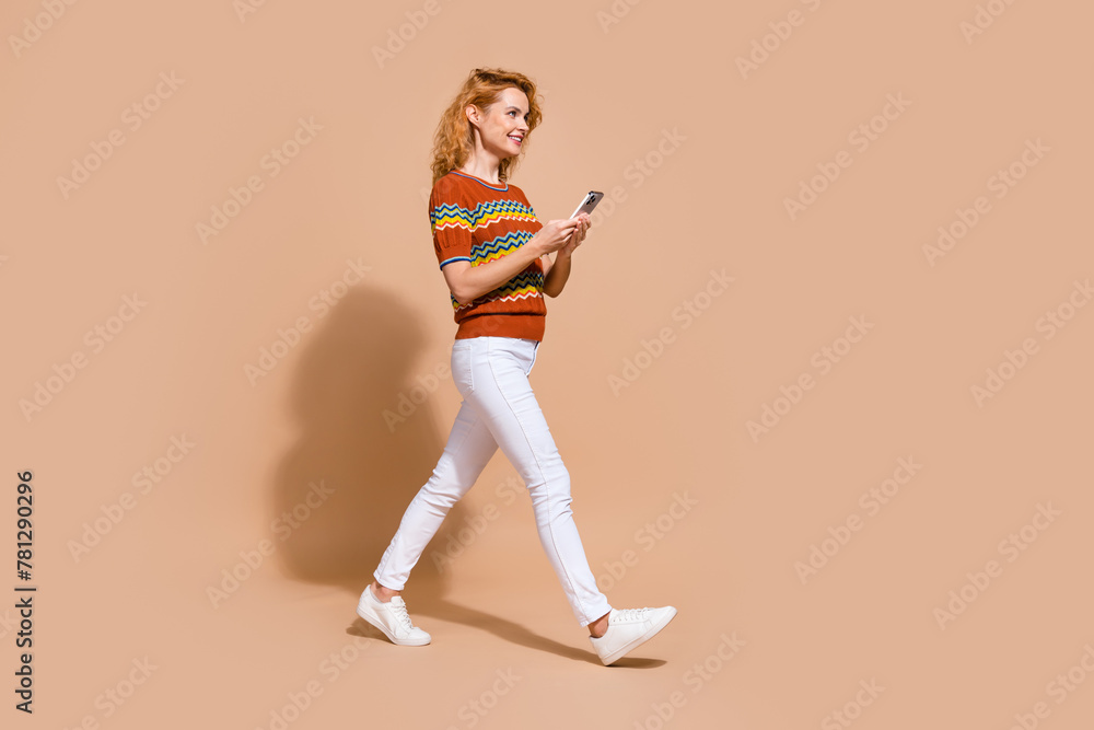 Full length photo of charming woman dressed knit t-shirt holding smartphone go look empty space isolated on pastel color background Full length photo of charming woman dressed knit t-shirt holding smartphone go look empty space isolated on pastel color background