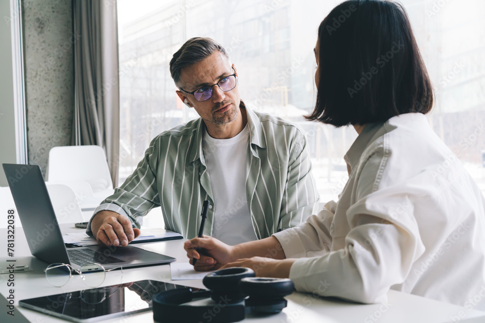 Fototapeta premium Male employee in optical spectacles for vision protection communicate with female colleague during cooperation process at table desktop with laptop computer, professional entrepreneurs talking