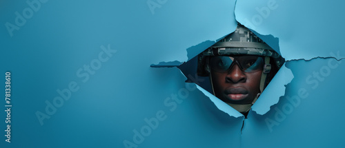 A focused soldier with night vision goggles peers through a tear in blue paper photo