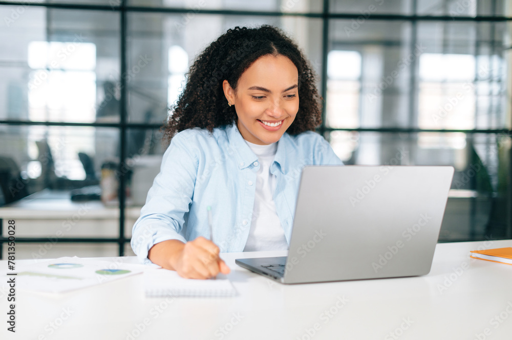 Happy successful brazilian or hispanic woman in a pastel blue shirt, company seo manager, sits in a modern office at work desk, working on a laptop, takes notes in notebook, develop project, smiles Happy successful brazilian or hispanic woman in a pastel blue shirt, company seo manager, sits in a modern office at work desk, working on a laptop, takes notes in notebook, develop project, smiles