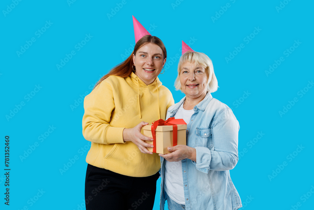 Young smiling happy daughter and mother together gifting birthday present with red ribbon isolated on blue color background studio. Human emotions, facial expression and holiday concept.