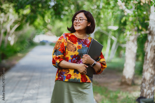 A smiling Asian woman with glasses holding a laptop in a sunny park. Happy entrepreneur enjoying remote work amidst green trees outdoors