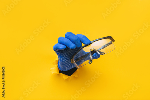 The right male hand in a blue fabric work glove holds safety glasses. Torn hole in yellow paper. Good job, eye protection and safety concept. Copy space.