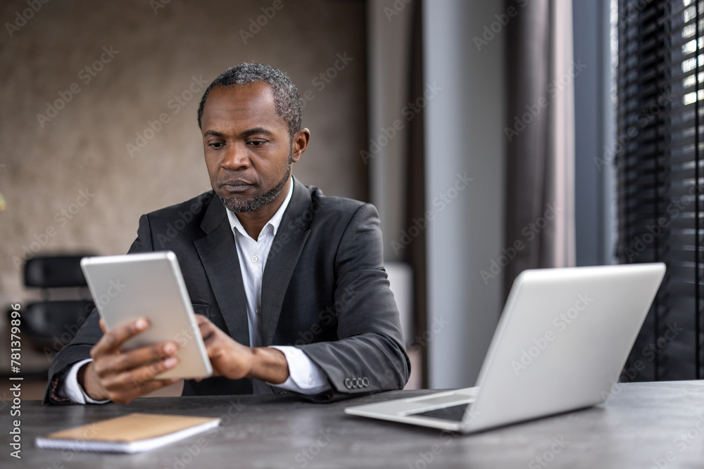 A man in a suit is sitting at a desk with a tablet and a laptop. He is focused on the tablet, possibly checking emails or browsing the internet. Concept of productivity and concentration A man in a suit is sitting at a desk with a tablet and a laptop. He is focused on the tablet, possibly checking emails or browsing the internet. Concept of productivity and concentration