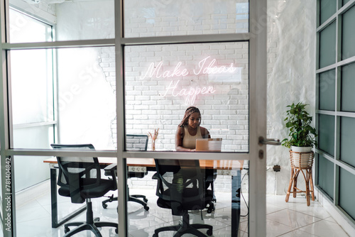 Concentrated African businesswoman working on laptop in chic office with motivational 'Make Ideas Happen' neon sign, symbolizing ambition and innovation © BullRun