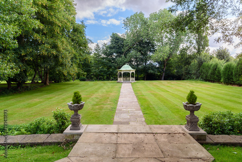 Deserted gazebo at the far end of a stone path through a lawn surrounded by trees in a park on partly cloudy summer day