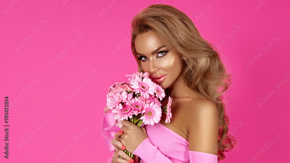 Elegant blonde woman wearing a fashionable pink dress holds spring flowers on a pink background.