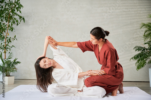 Masseuse making thai yoga massage. Woman therapist doing traditional massage treatment, stretching female patient's body. Client sitting on mat, enjoying hand massage in calm atmosphere. photo