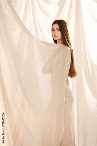 A young woman with long brunette hair posing in front of a white curtain, exuding a summer mood in her stylish outfit. © LIGHTFIELD STUDIOS