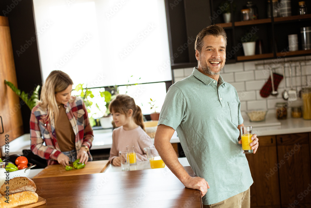 Joyful man with a glass of orange juice while a woman and child prepare food in a cozy kitchen Joyful man with a glass of orange juice while a woman and child prepare food in a cozy kitchen