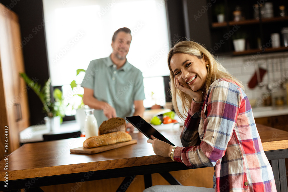 A woman reads from a tablet at the kitchen counter while a man holding an tomatoes smiles at her
