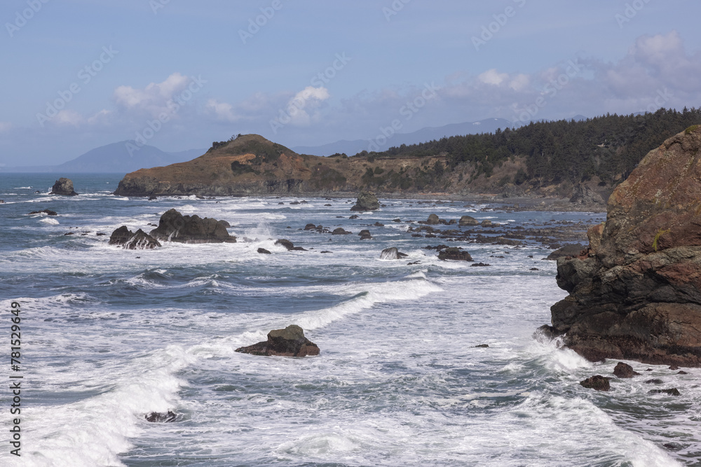 Obraz premium The ocean is rough and the rocks are scattered throughout the water. The waves are crashing against the shore, creating a powerful and dynamic scene. The rocky shoreline adds a sense of ruggedness