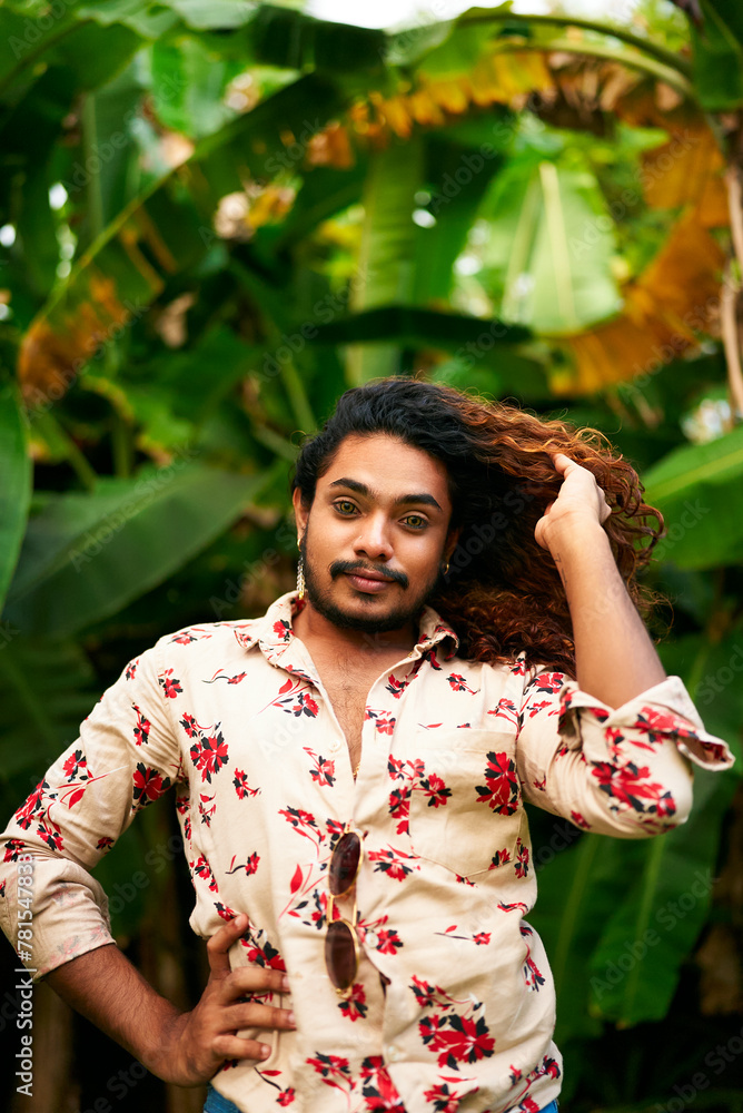 Vibrant man with rich curly hair, floral shirt stands in tropical green plants. He poses confidently, hand in hair, bold fashion, expression of LGBTQ pride and joy in nature backdrop.