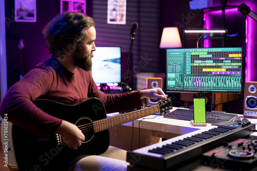 Musician learning to tune his guitar with internet tutorial via greenscreen on phone, twisting the knobs and practicing music lessons. Artist songwriter does rehearsal with acoustic instrument.