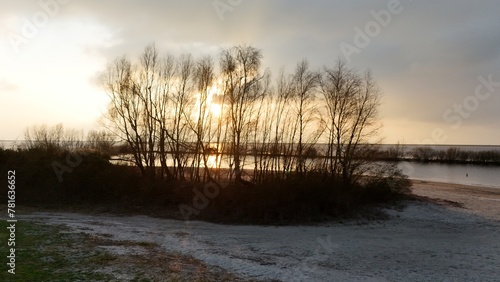 Fototapeta Naklejka Na Ścianę i Meble -  Sunset over a tranquil lake with silhouettes of trees and a light dusting of snow on the ground, reflecting a serene winter evening.