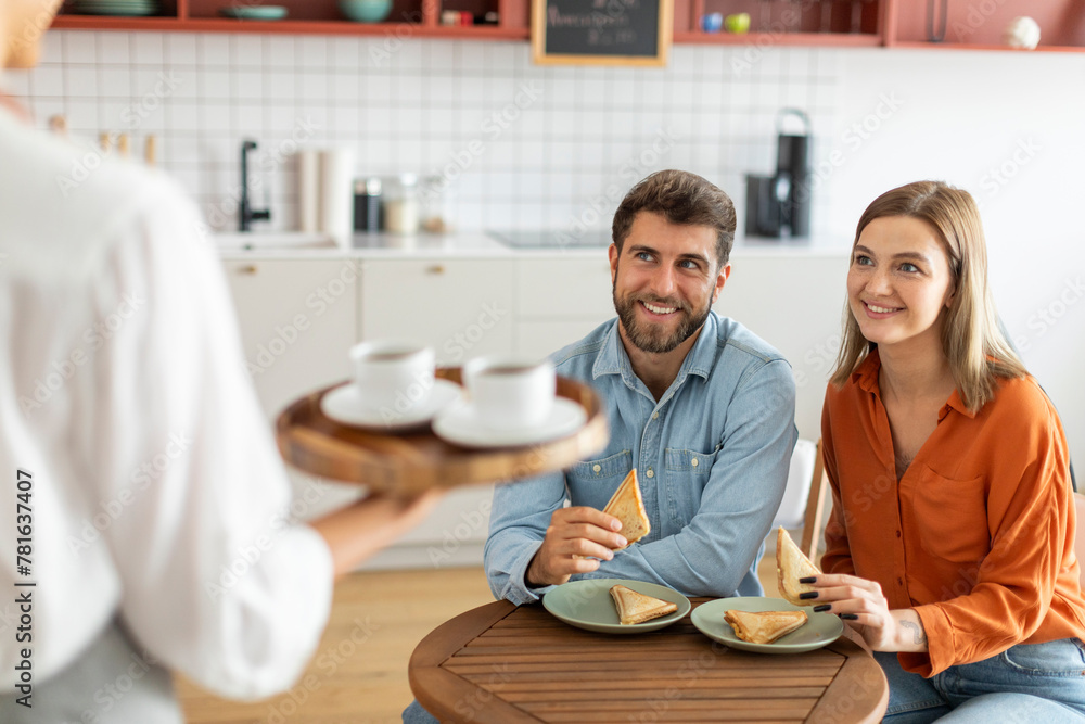 Waitress serving hot aromatic brewed coffee drinks to young couple, holding tray with cups, man and woman visitors waiting at cafe table Waitress serving hot aromatic brewed coffee drinks to young couple, holding tray with cups, man and woman visitors waiting at cafe table