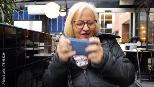 blonde gamer grandmother with glasses playing on the smartphone in a cafe loses the game and laughs in slow motion photo