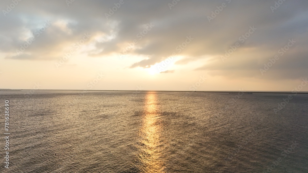 Obraz premium Sunset over calm sea with clouds partially covering the sun, casting a golden reflection on the water.