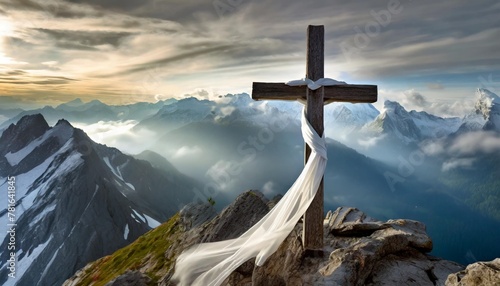 white satin scarf tied around weathered wooden cross on top of a windswept mountain peak overlooking a range of jagged rocky peaks partially obscured by mist photo