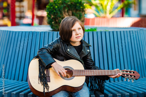 A beautiful little girl, in rock style,in a leather jacket and jeans,sits and plays an acoustic guitar