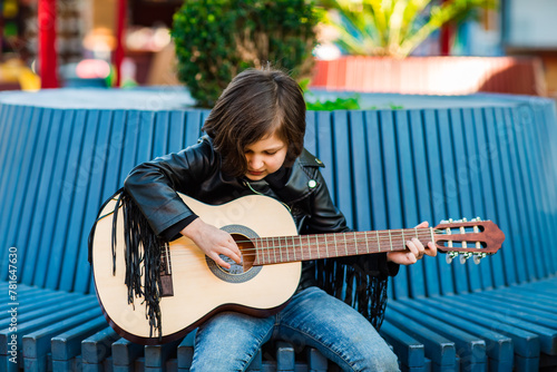 A beautiful little girl, in rock style,in a leather jacket and jeans,sits and plays an acoustic guitar