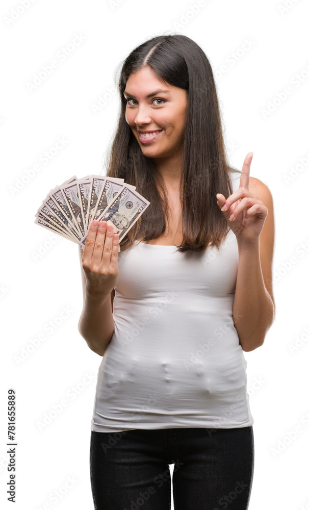 Young hispanic woman holding dollars surprised with an idea or question pointing finger with happy face, number one Young hispanic woman holding dollars surprised with an idea or question pointing finger with happy face, number one