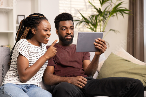 Couple Enjoying Digital Tablet Technology Together At Home. Smiling Young Woman Pointing At Screen While Relaxed Man Holds Tablet. Indoor Leisure, Loving Relationships, And Modern Lifestyle Concept. 