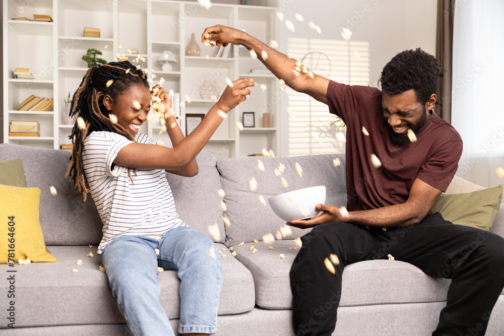 Joyful Couple Having Fun With Popcorn On Couch. African American Man And Woman Enjoying Playful Time, Home Entertainment. Lifestyle, Leisure, Togetherness Concept Captured.  - obrazy, fototapety, plakaty 