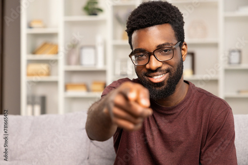 Smiling African American Man Pointing At Camera In Modern Living Room. Selective Focus On Face With Blurry Bookshelf Background. Concept Of Invitation, Engagement, And Direct Communication